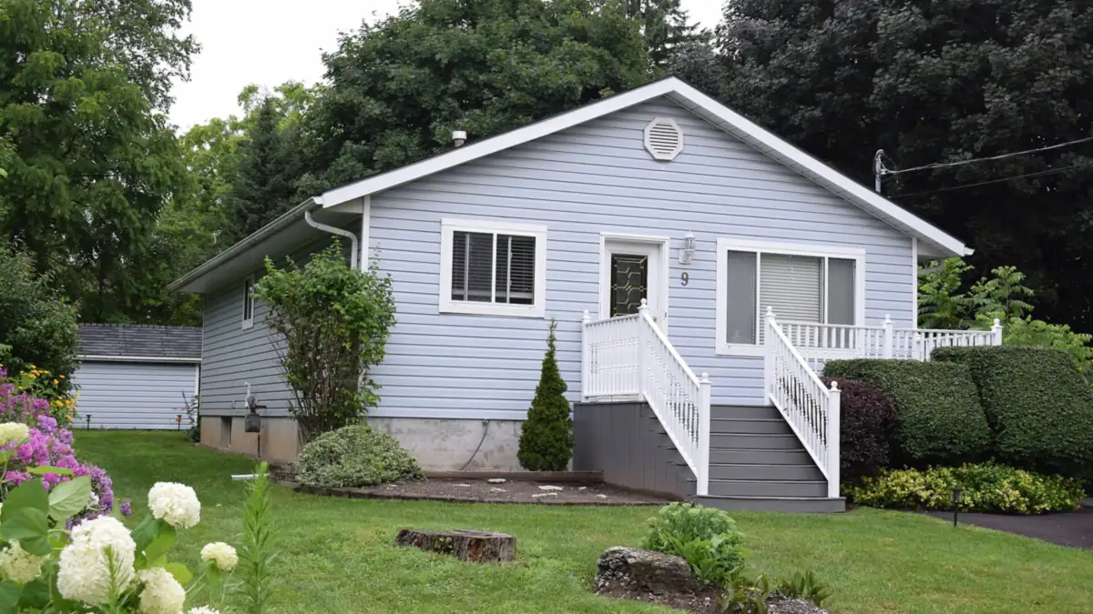 Pale blue bungalow with white porch railings, trimmed hedges and a green lawn