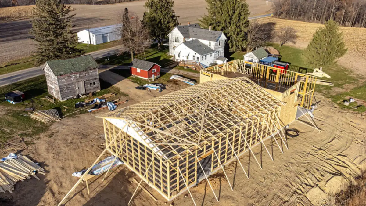Timber-framed building under construction beside a farmhouse, seen from above
