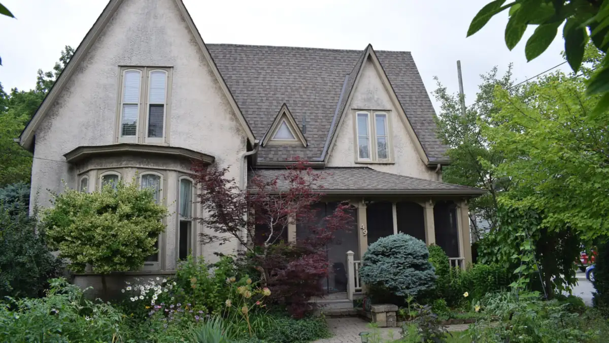 Gabled stucco house with a bay window and screened porch behind a leafy garden