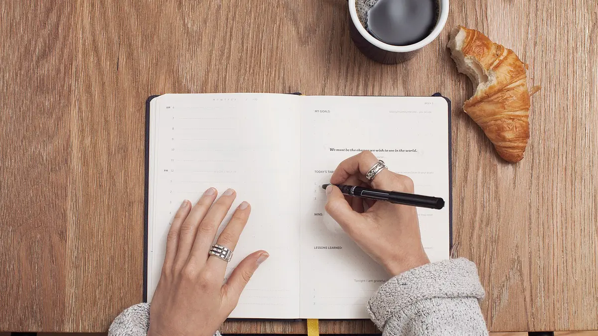 Hands writing in an open planner on a wooden table beside coffee and a croissant