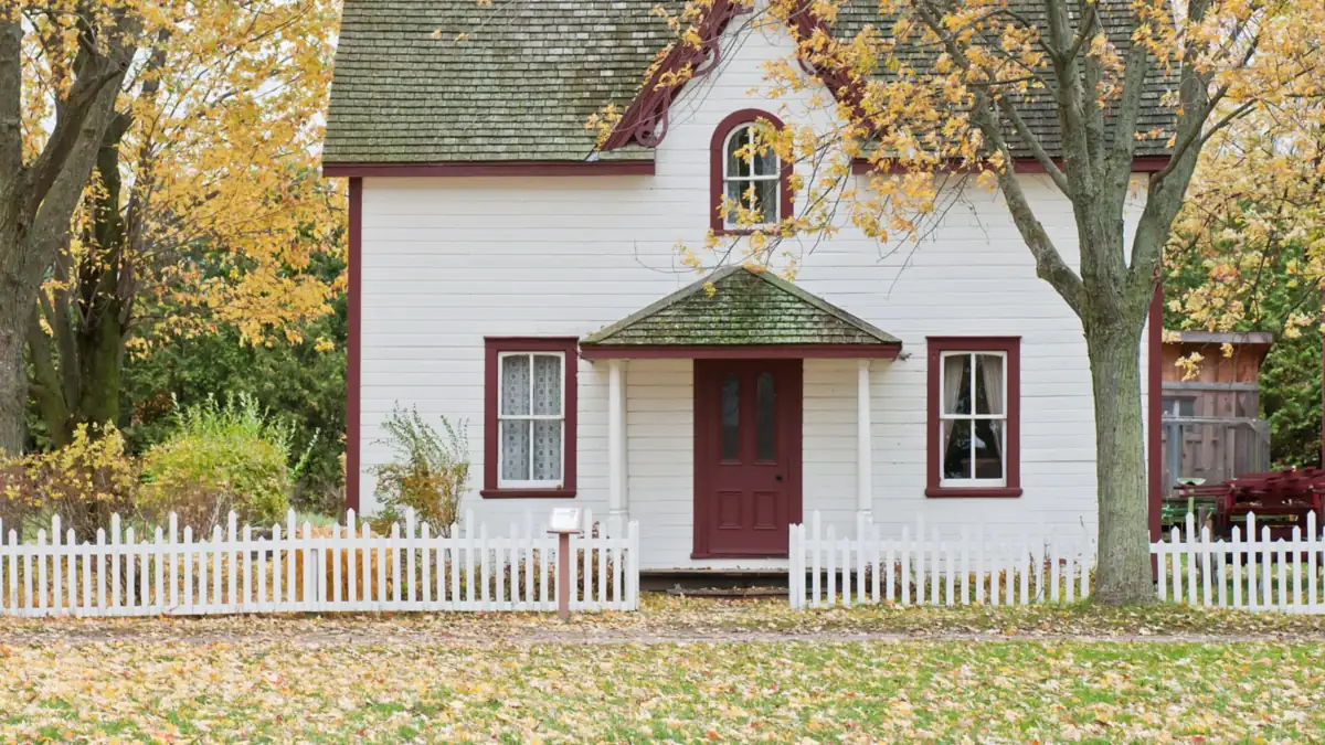 Small white house with a red door and picket fence under yellow autumn leaves