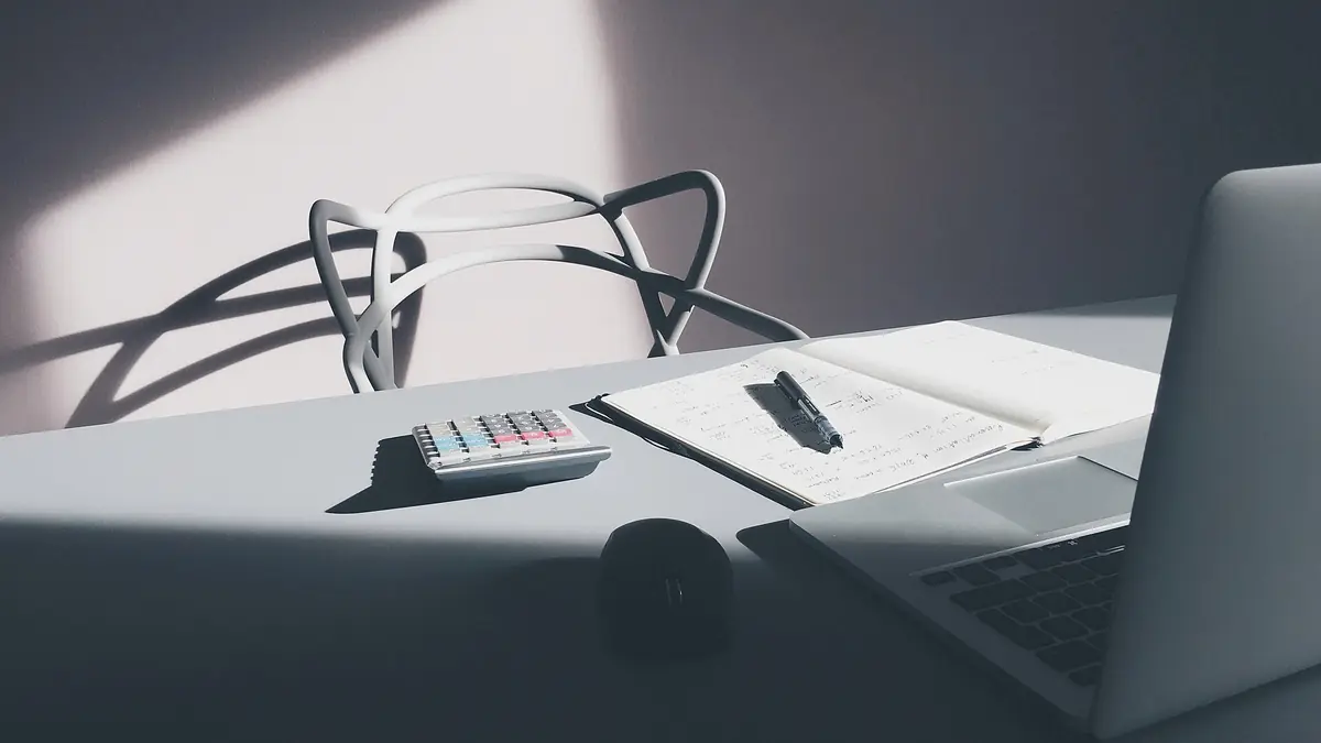 Desk with a calculator, an open notebook and pen, a mouse and a laptop in window light