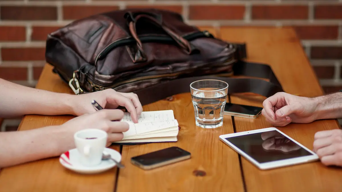 Two people talking across a wooden table with a notebook, coffee and a tablet