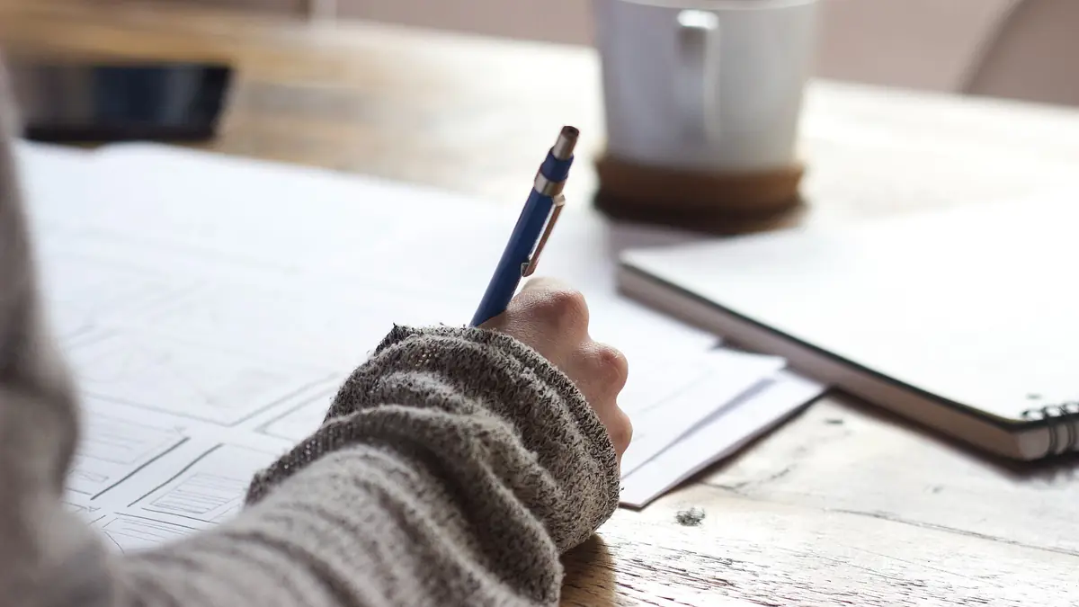 Person writing on printed paperwork at a wooden table beside a white mug