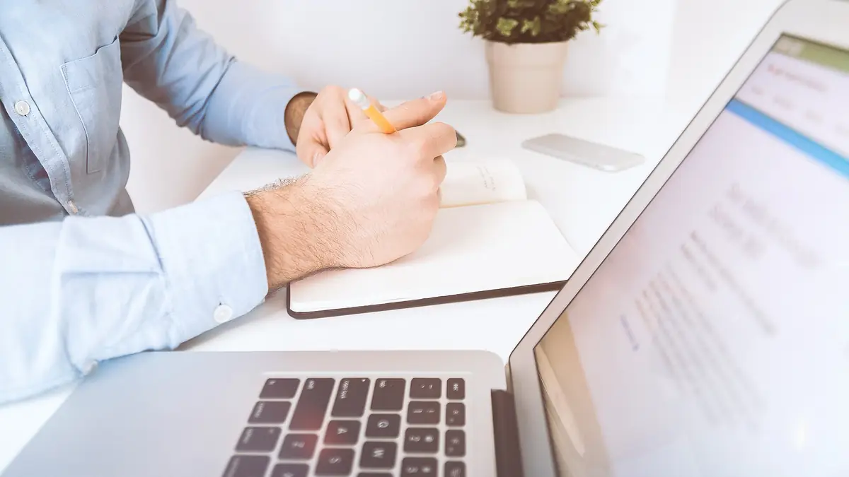 Person in a blue shirt writing notes in a notebook beside an open laptop